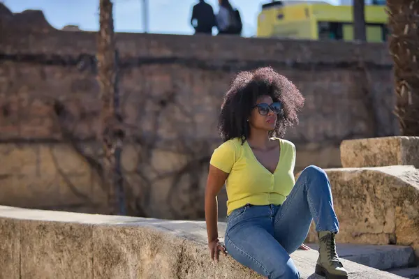 美しい アフロの髪を持つ黒の女性は 黄色のTシャツ ジーンズやサングラスを着て 石の壁に座って リラックスし 穏やかな日光浴 休暇の概念 — ストック写真