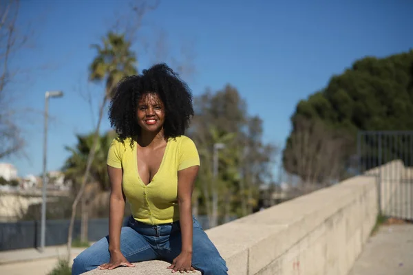 黄色いTシャツとジーンズを着て 石の壁に座って 笑顔で幸せでセクシーな若い 美しい黒人女性 コンセプト — ストック写真
