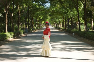 Bej renkli çingene flamenko takım elbiseli ve kırmızı şallı genç siyah ve Güney Amerikalı kadın, İspanya 'nın Seville şehrinde bir parkta poz veriyor. Konsept dansı, folklor, flamenko, sanat.