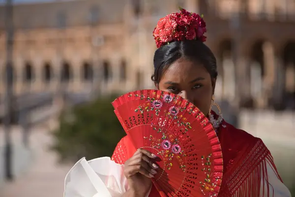 Portrait of a young black and South American woman in a beige flamenco gypsy costume and red shawl, covering herself from the sun with a fan in Seville in Spain. Concept dance, folklore, flamenco, art