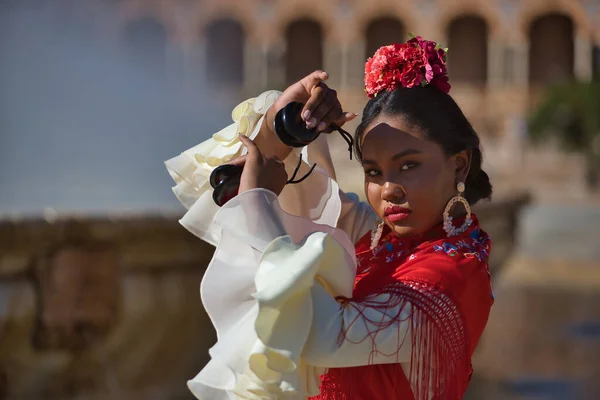 Young black and South American woman in a beige gypsy flamenco suit and red shawl, dancing with castanets in a beautiful square in the city of Seville in Spain. Concept dance, folklore, flamenco, art