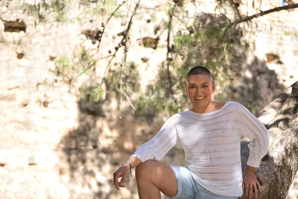 Portrait of non-binary person, young and South American, heavily makeup, leaning on a tree trunk, smiling very happy, looking at camera. Concept queen, lgbtq+, pride, queer.