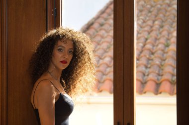 Genç bir kadın, güzel, esmer, kıvırcık saçlı, siyah elbiseli, kameraya bakan rahat, sakin ve kendinden emin. Moda, güzellik, güven kavramı.