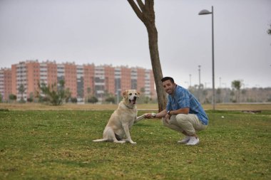 Genç İspanyol adam, çimenlerde çömelmiş köpeğinin patisini tutuyor, kameraya bakıyor. Kavram, köpekler, hayvanlar, hayvanlar, arkadaşlar.