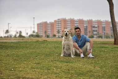 Genç İspanyol adam, köpeğinin yanındaki çimlerin üzerinde oturuyor, ikisi de kameraya bakıyor. Kavram, köpekler, hayvanlar, hayvanlar, arkadaşlar.