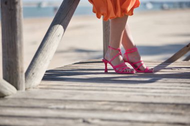 Yüksek topuklu, pembe kayışlı, sahilde tahta bir yolda yürüyen kadınların ayakları. Kavramsal ayak, moda, ayakkabı.