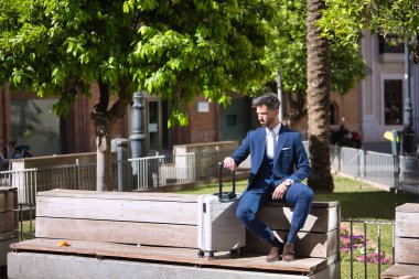 Sakallı ve takım elbiseli genç ve çekici bir işadamı, bavulunun yanındaki bankta oturmuş taksi bekliyor. Güzelliği, modayı, başarıyı, başarıyı, eğilimi kavra.