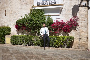 Sakallı, takım elbiseli ve kravatlı çekici genç bir işadamı, çiçek arkaplanlı elleri ceplerinde poz veriyor. Güzelliği, modayı, başarıyı, başarıyı, eğilimi kavra.