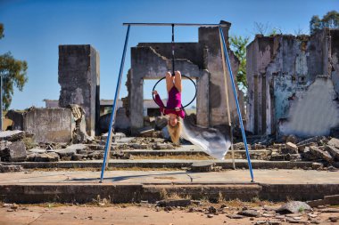 Genç bir kadın, sarışın ve atletik. Havada asılı duran bir halkayla baş aşağı egzersiz yapıyor. Rüzgarda savrulan beyaz bir duvak, harabeler içindeki bazı binaların yanında..