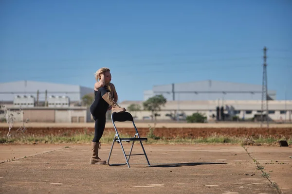 Young woman, blonde and athletic, in a black body and military boots, thoughtful, resting her foot on an outdoor chair. Concept thoughts, dreams, worry.