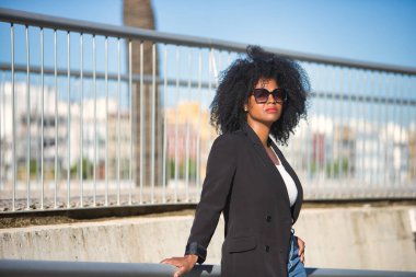 Genç, güzel, siyah saçlı, ceketli ve güneş gözlüklü metal korkuluklara yaslanmış, sakin ve rahat güneş ışınları alan bir kadın. Barış, sükunet, güncel, modern kavramları.