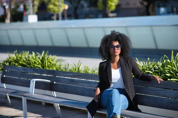 stock image Young, beautiful, black woman with afro hair, with jacket and sunglasses sitting on a bench, receiving the sun's rays, relaxed and calm. Relaxed, calm, modern concept.