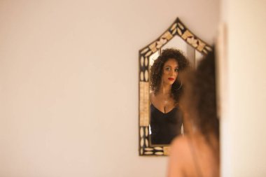 Genç bir kadın, güzel, esmer, kıvırcık saçlı, siyah elbiseli, aynaya bakan, kendini bulan. Moda, güzellik, yansımalar, ayna, içgözlem.