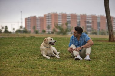 Genç İspanyol bir adam, köpeğinin yanındaki çimlerin üzerinde oturuyor ve sanki konuşuyorlarmış gibi birbirlerine bakıyor. Kavram, köpekler, hayvanlar, hayvanlar, arkadaşlar.