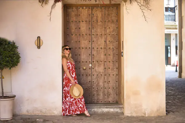 Güney Amerikalı kadın, genç, güzel, çiçek desenli elbiseli, güneş gözlüklü ve şapkalı, ahşap kapı çerçevesine yaslanmış, rahat ve sakin. Konsept seyahat, varış yerleri, Avrupa, Akdeniz, tatil
