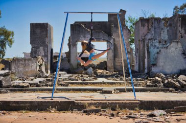 Genç, sarışın, atletik bir kadın. Havada asılı duran bir halkayla bazı yıkık binaların yanında egzersiz yapıyor. Kavramsal çember, yoga ve direk dansı.