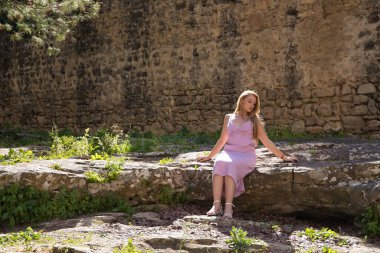 Young, beautiful, blonde woman in a pink dress, sitting on a stone inside an ancient medieval castle receiving the rays of the sun, alone, quiet and peaceful. Concept beauty, relax, tranquility, calm
