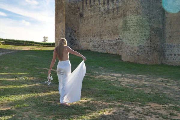 Genç bir kadın, güzel ve sarışın, beyaz elbiseli, onu eliyle kaldırıyor, diğerinde ayakkabıları, sırtı şatonun yanında, güneşin alevleri, sakin ve rahat..