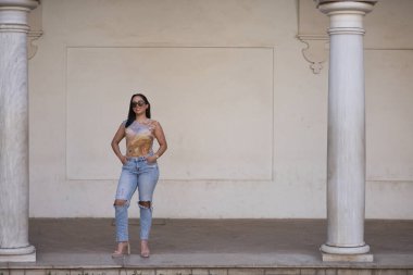 Genç, güzel, esmer, desenli tişörtlü ve yırtık kotlu, elleri ceplerinde gülümserken poz veren Güney Amerikalı bir kadın. Güzelliği, modayı, eğilimi kavra.