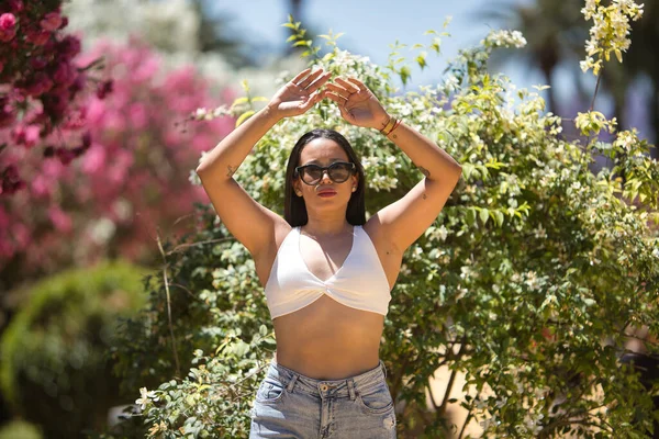 stock image Young, beautiful, brunette and South American woman with white top, torn jeans and sunglasses, with her hands up, receiving the sun's rays in the middle of a park. Concept beauty, peace, tranquility.