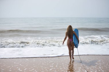 Genç, güzel, sarışın bir kadın, pembe bir bikini giyiyor, kolunun altında, sırtında mavi bir sörf tahtası var, denizin dalgalarına doğru koşuyor, sahilden. Kumsal, bikini, sörf, dalgalar, spor.