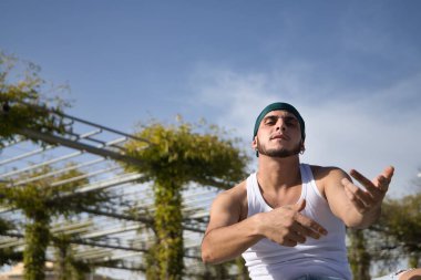 Latino and Hispanic boy, young, rebellious, wearing headscarf, looking defiantly at camera and gesturing as if rapping, sitting on a bench. Troubled concept, rebel, gangs, rapping, defiant.