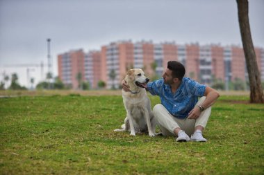 Genç İspanyol adam, çimlerin üzerinde oturuyor köpeğine sarılıyor ve ona şefkatle bakıyor. Kavram, köpekler, hayvanlar, hayvanlar, arkadaşlar.
