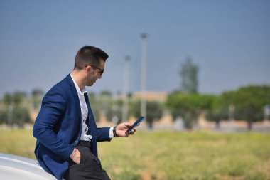 Başarılı, çekici, ceketli ve güneş gözlüklü İspanyol genç adam beyaz lüks arabasının kaputuna yaslanmış cep telefonuna danışıyor. Kavram, arabalar, lüks, başarı, iş adamı.