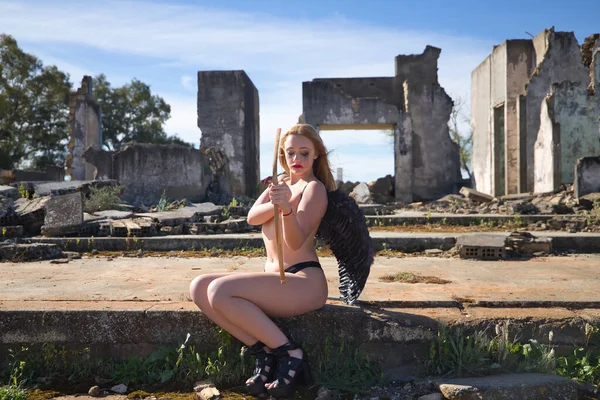 Young, beautiful, blonde woman with black angel wings, with the paint on her eyes smeared, with bow and arrow, sitting in the middle of some ruined buildings. Concept fallen angel, lack of love.