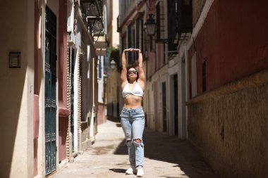 Genç bir kadın, güzel, esmer, beyaz bluzlu, yırtık kot pantolon ve güneş gözlüklü, kolları havada, ıssız bir sokakta güneş ışınlarını alan. Güzelliği, modayı, barışı, huzuru kavra.