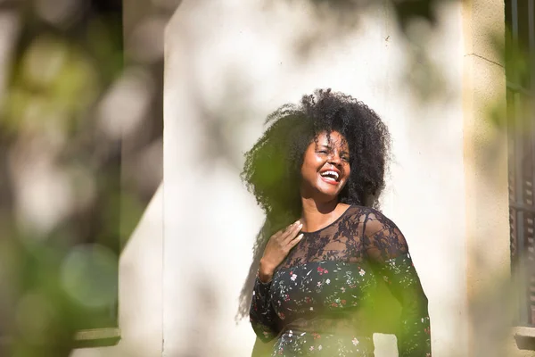 Young woman, beautiful and black with afro hair, with flower dress leaning on a wall, very smiling and happy, seen through a green plant. Concept beauty, laughter, happiness, fun, plants.
