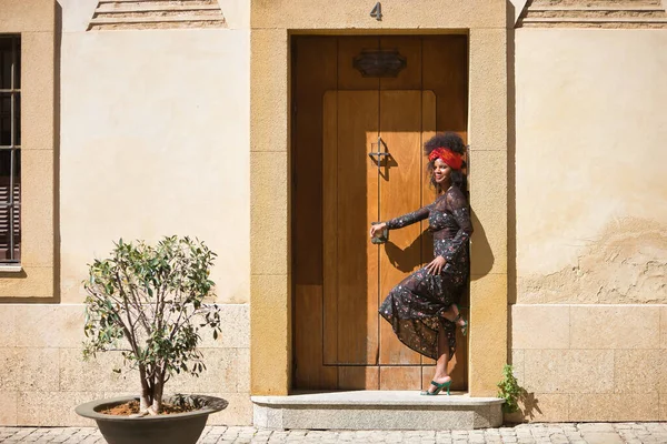 Young woman, beautiful and black with afro hair, with flower dress and scarf in her hair, leaning on the frame of a wooden door in a fun and playful attitude. Concept beauty, ethnicity, fun, play.