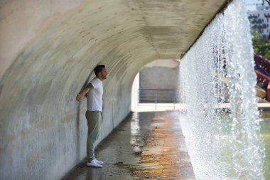 Genç, çekici ve İspanyol bir adam, şelalenin olduğu bir köprünün altındaki duvara yaslanıyor. Kavram, güzellik, su, erkeklik.