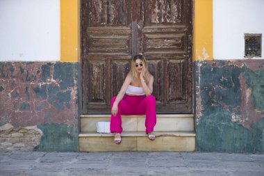 Güney Amerikalı bir kadın, genç, güzel ve sarışın, beyaz bluzlu, pembe pantolonlu ve güneş gözlüklü, tahtadan bir kapının pencere eşiğinde otururken, şehvetli ve kışkırtıcı bir tavırla poz veriyor..