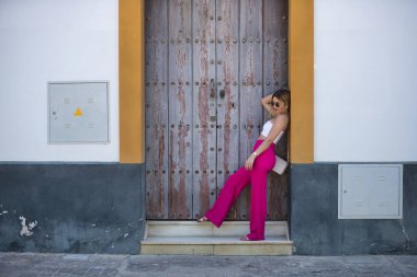 Güney Amerikalı bir kadın, genç, güzel ve sarışın, beyaz bluzlu, pembe pantolonlu ve güneş gözlüklü, ahşap bir kapının çerçevesine yaslanmış poz veriyor. Trend konsepti, güzellik, moda.