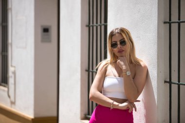 Güney Amerikalı bir kadın, genç, güzel ve sarışın, beyaz bluzlu, pembe pantolonlu ve güneş gözlüklü, beyaz bir duvara yaslanmış, rahat ve sakin bir tavırla poz veriyor. Kavram eğilimi, güzellik, moda.