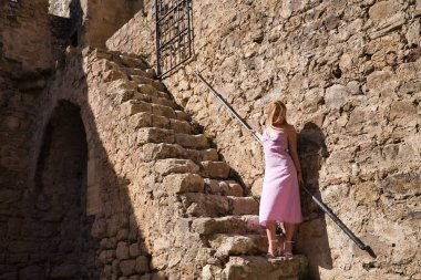 Genç, güzel, pembe elbiseli sarışın kadın, eski bir ortaçağ şatosunun korkuluklarına tutunuyor. Güzelliği, modayı, merdivenleri, kaleleri kavra.