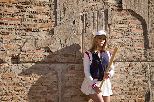Young, pretty, blonde woman in white cap and skirt, jacket and baseball bat leaning against broken brick wall in background. Concept beauty, sport, baseball, bat, ball, glove.