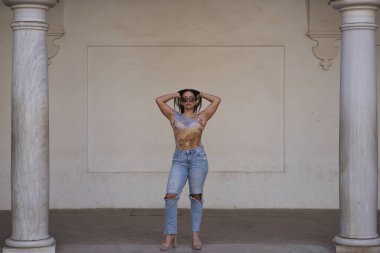 Genç, güzel, esmer ve baskılı tişörtü ve yırtık kotu olan, elleri başının üstünde, bağımsız ve güçlü poz veren Güney Amerikalı bir kadın. Güzelliği, modayı, eğilimi, bağımsızlığı, güçlendirmeyi kavra.