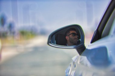 Başarılı, çekici, ceketli ve güneş gözlüklü genç İspanyol adam beyaz lüks arabasının aynasında yansıyor. Kavram, arabalar, lüks, başarı, iş adamı.