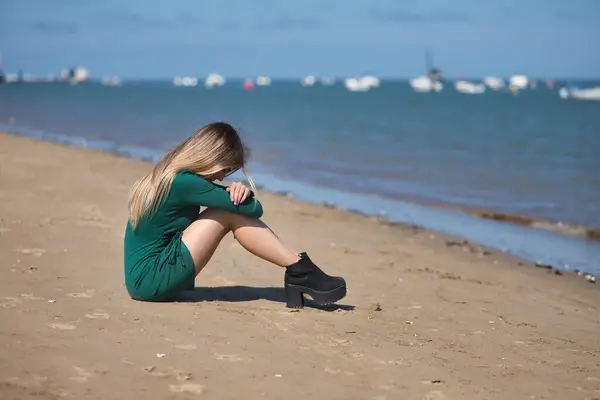 Genç, güzel, sarışın bir kadın, yeşil bir elbise giyiyor, plajın kıyısında oturuyor, kafası dizlerinin arasında ve bacaklarını sıkıca tutuyor, ağlıyor, üzgün ve depresif. Depresyonu, üzüntüyü kabul et.