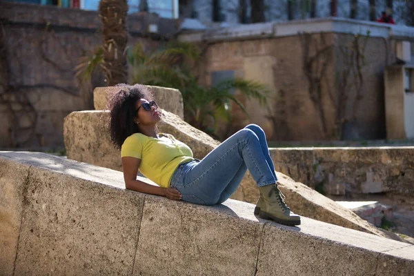 Young, beautiful, black woman with afro hair, wearing a yellow t-shirt, jeans and sunglasses, lying on a stone wall, relaxed, calm and sunbathing. Vacation concept, travel, current, modern.