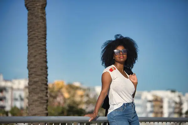 Young, beautiful, black woman with afro hair, white t-shirt and sunglasses leaning on a metal railing, on vacation, receiving the sun\'s rays. Concept travel, relax, vacation, current, modern.