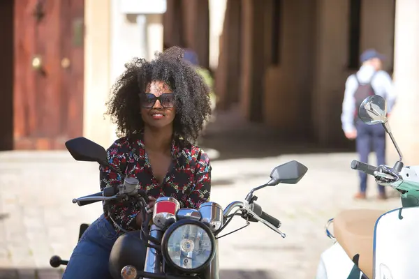 Young woman, beautiful and black with afro hair, with floral shirt and sunglasses, mounted on a motorcycle in a happy and fun attitude. Motorcycle concept, fun, transport.