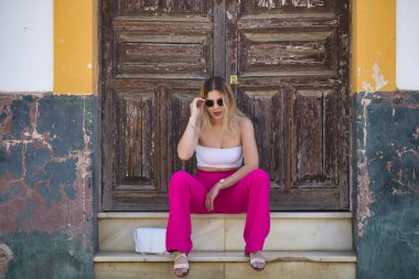 Güney Amerikalı bir kadın, genç, güzel ve sarışın, beyaz bluzlu, pembe pantolonlu ve güneş gözlüklü, tahtadan bir kapının pencere eşiğinde otururken, şehvetli ve kışkırtıcı bir tavırla poz veriyor..