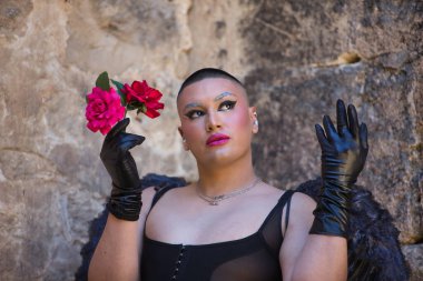 İkili olmayan bir kişinin portresi, genç ve Güney Amerikalı, çok makyajlı, siyah kanatlı ve deri eldivenli, korse, elinde güllerle poz veriyor. Kavram kraliçesi, lgbtq +, gurur, nonoş.