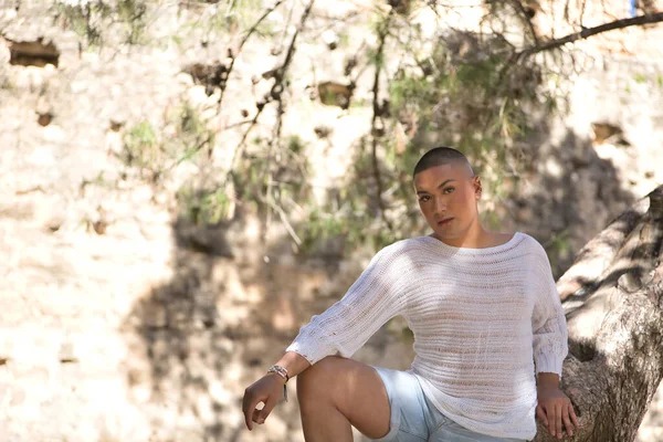 Portrait of non-binary person, young and South American, heavily makeup, leaning on a tree trunk, relaxed, looking at camera. Concept queen, lgbtq+, pride, queer.