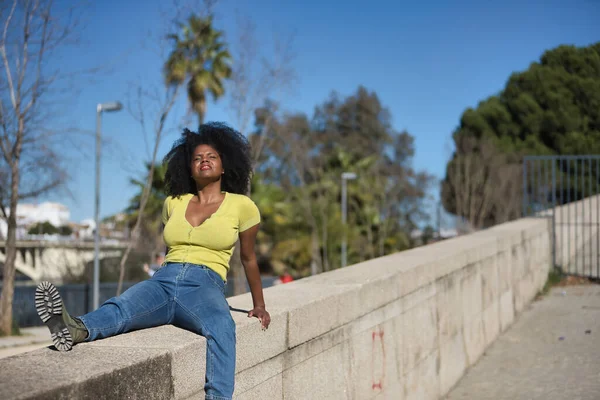 Young, beautiful, black woman with afro hair, wearing a yellow t-shirt, jeans and boots, sitting on a stone wall, relaxed and calm, sunbathing. Concept of relaxation, tranquility, peace.