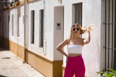 Güney Amerikalı kadın, genç, güzel ve sarışın, beyaz bluzlu, pembe pantolonlu ve güneş gözlüklü, saçlarına dokunurken poz veriyor. Kavram eğilimi, güzellik, moda.