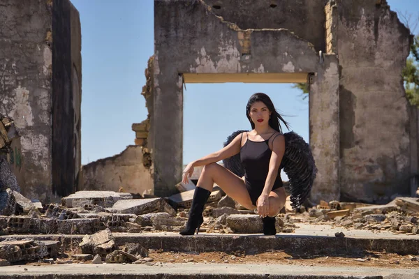 South American woman, young, beautiful, brunette with lingerie and black wings, posing crouched in the middle of ruined buildings relaxed and calm. Concept angels, beauty, costumes, wings, halloween.